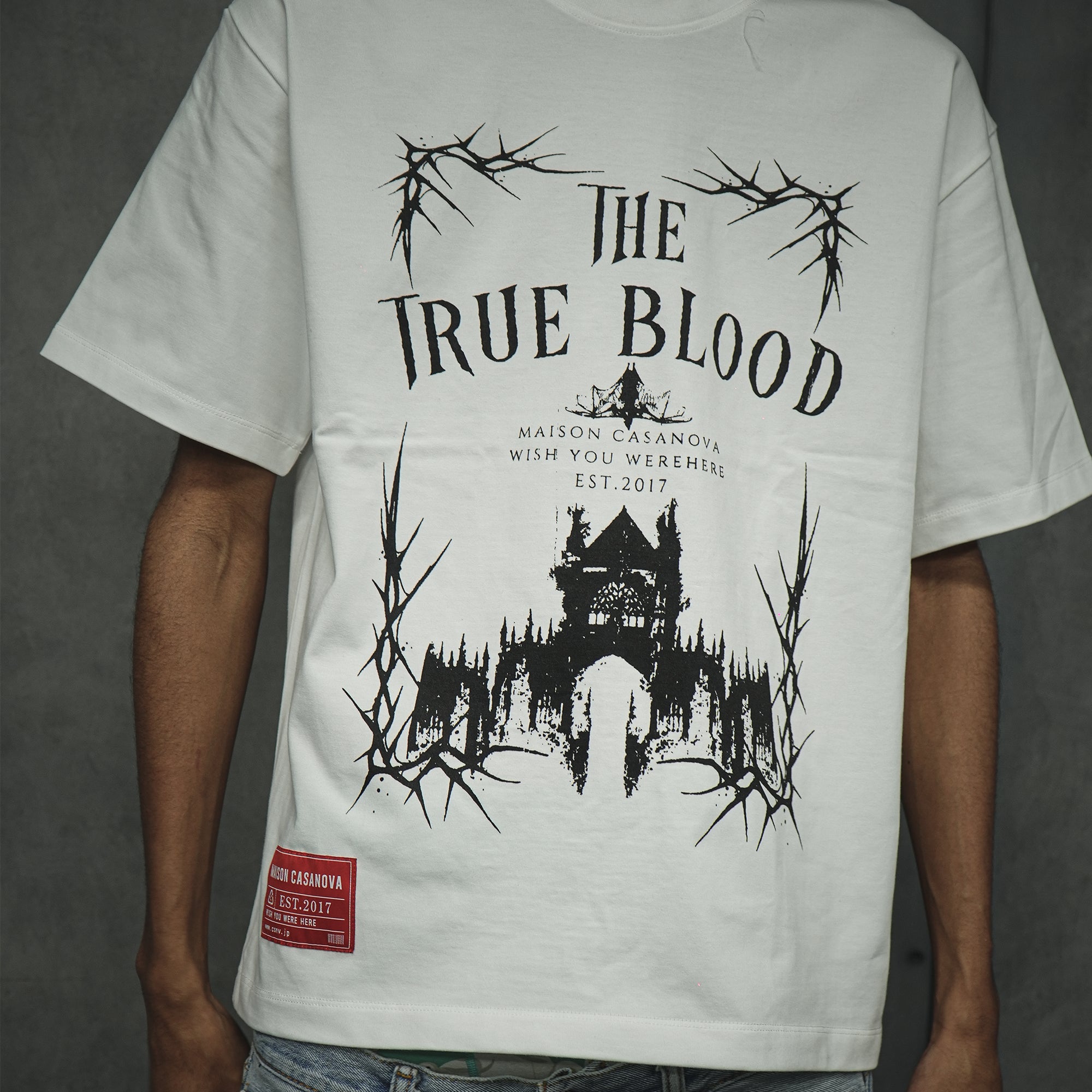 メゾンカサノバ 白のグラフィックTシャツ。フロントに「THE TRUE BLOOD」の文字とゴシック調の城、イバラの白いイラスト。左裾に赤いブランドラベル付き。